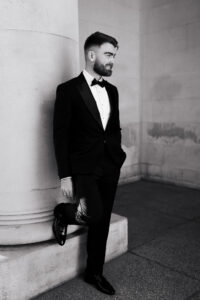 Black-and-white portrait of the groom with one leg on a pillar