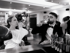 Black-and-white moment of the bride and groom drinking champagne at the bar